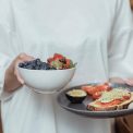 Person holding a bowl of fresh berries and a plate with avocado toast, displaying a healthy breakfast spread.