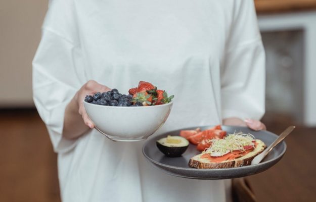 Person holding a bowl of fresh berries and a plate with avocado toast, displaying a healthy breakfast spread.