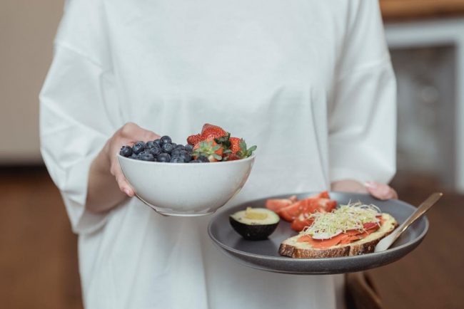 Person holding a bowl of fresh berries and a plate with avocado toast, displaying a healthy breakfast spread.